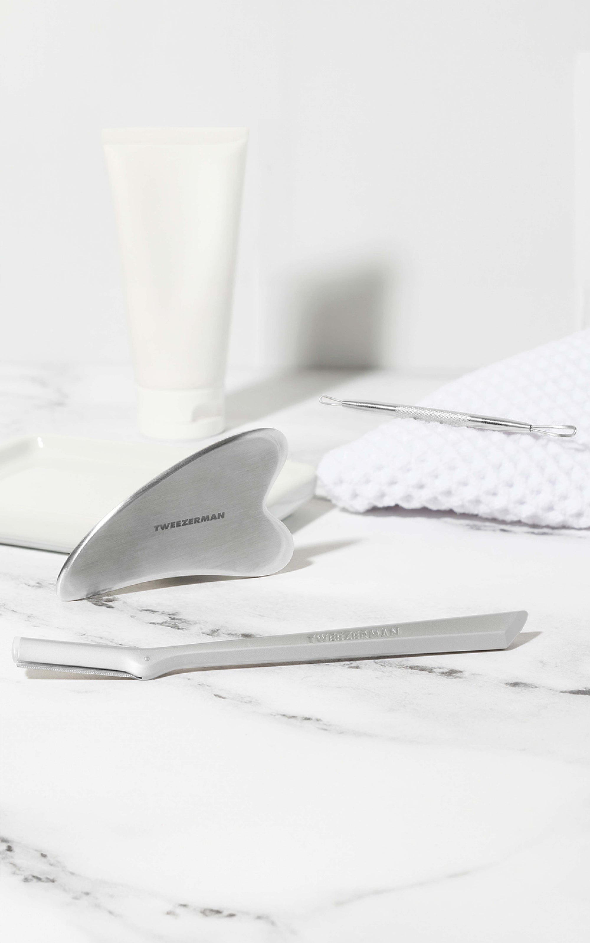 Tweezerman-Schönheitswerkzeuge arrangiert auf weißem Keramikobjekt und Handtuch auf einer weißen Mamorplatte.