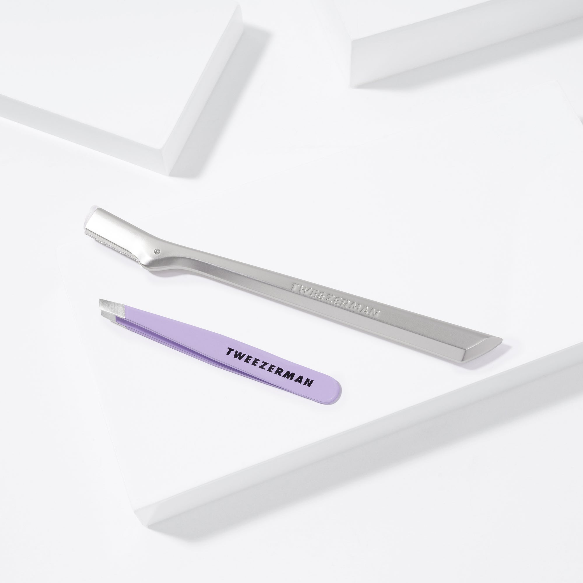 TWEEZERMAN Mini schräge Pinzette in lavender und Augenbrauenrasierer in silber auf weißen Podesten.