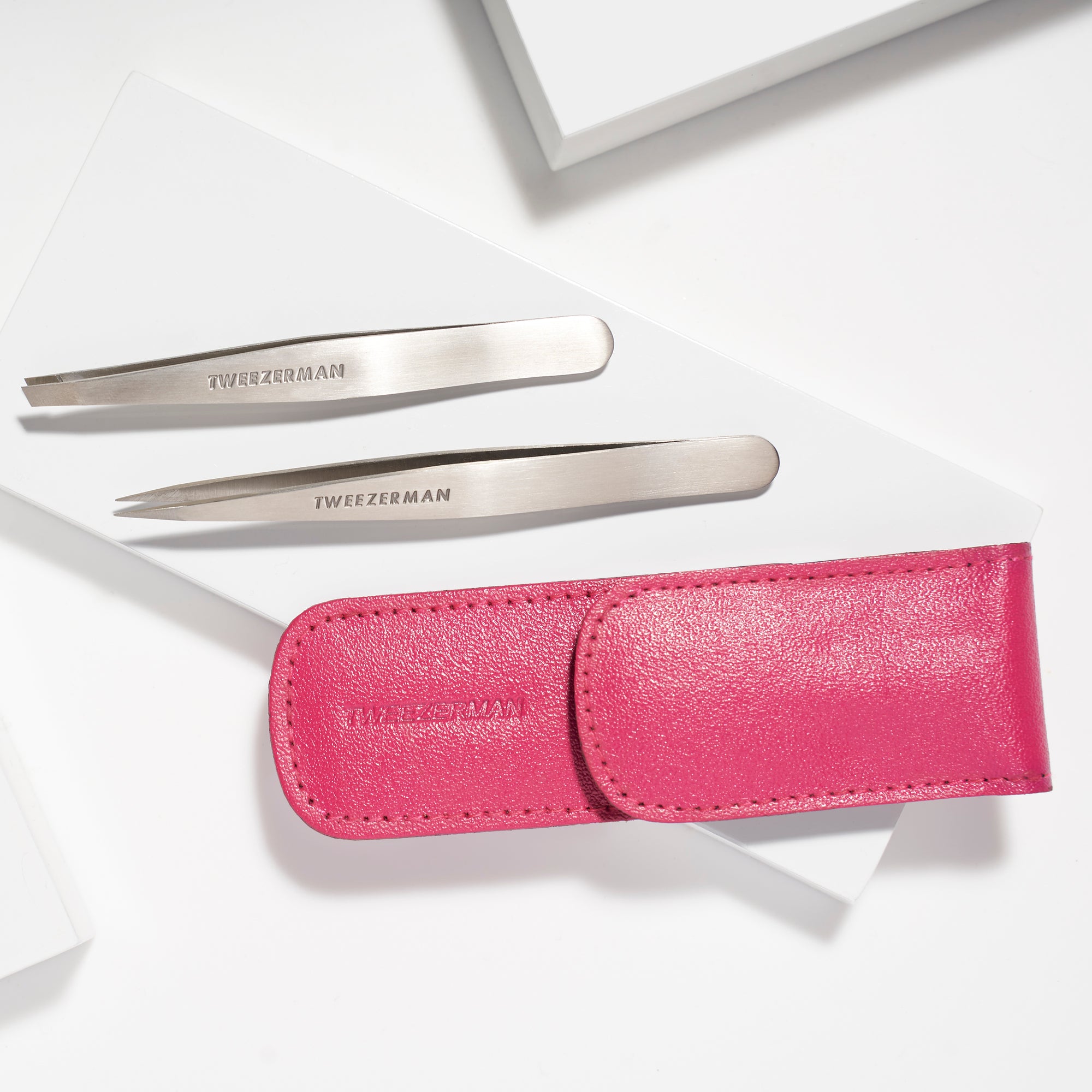 TWEEZERMAN Petite Set mit schräger und spitzer Pinzette in einem pinken Lederetui liegend auf weißen Podesten.