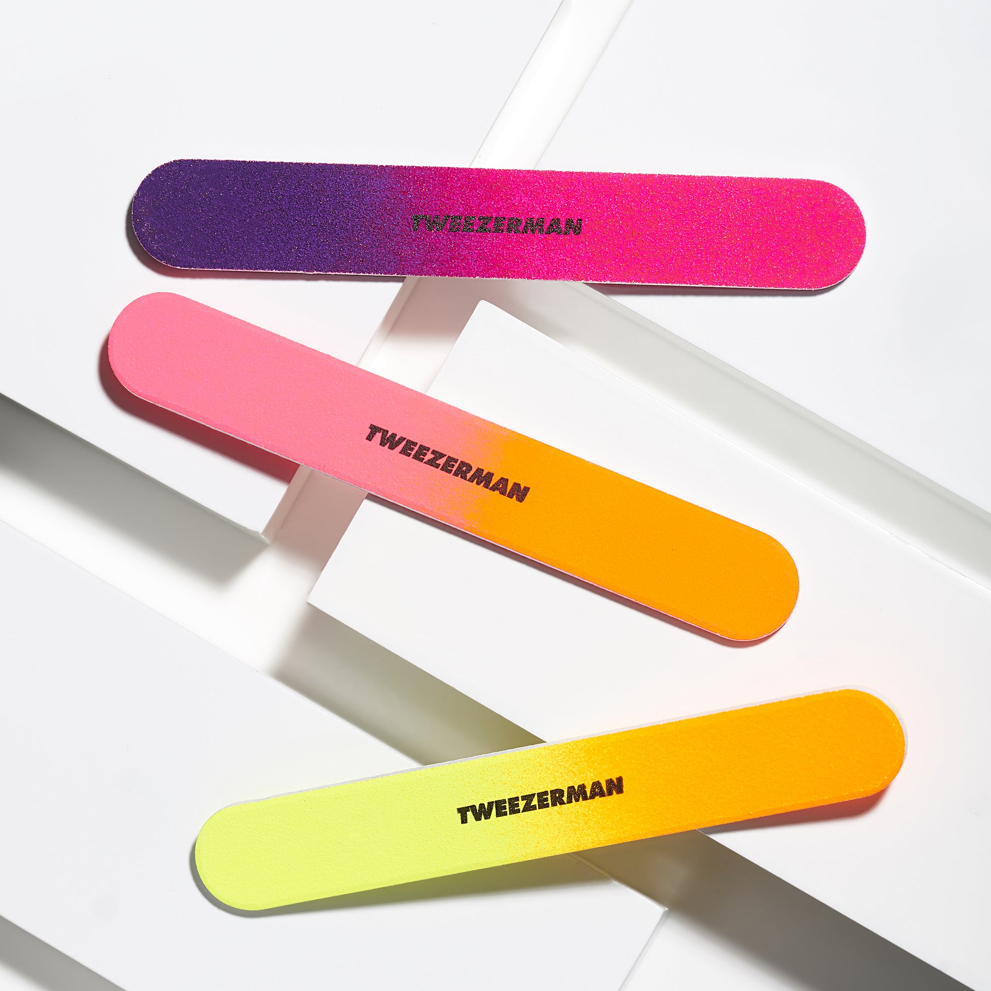 Drei TWEEZERMAN Nagelfeilen in verschiedenen Neonfarben, mit zwei unterschiedlichen Anwendungsflächen auf weißen Podesten.