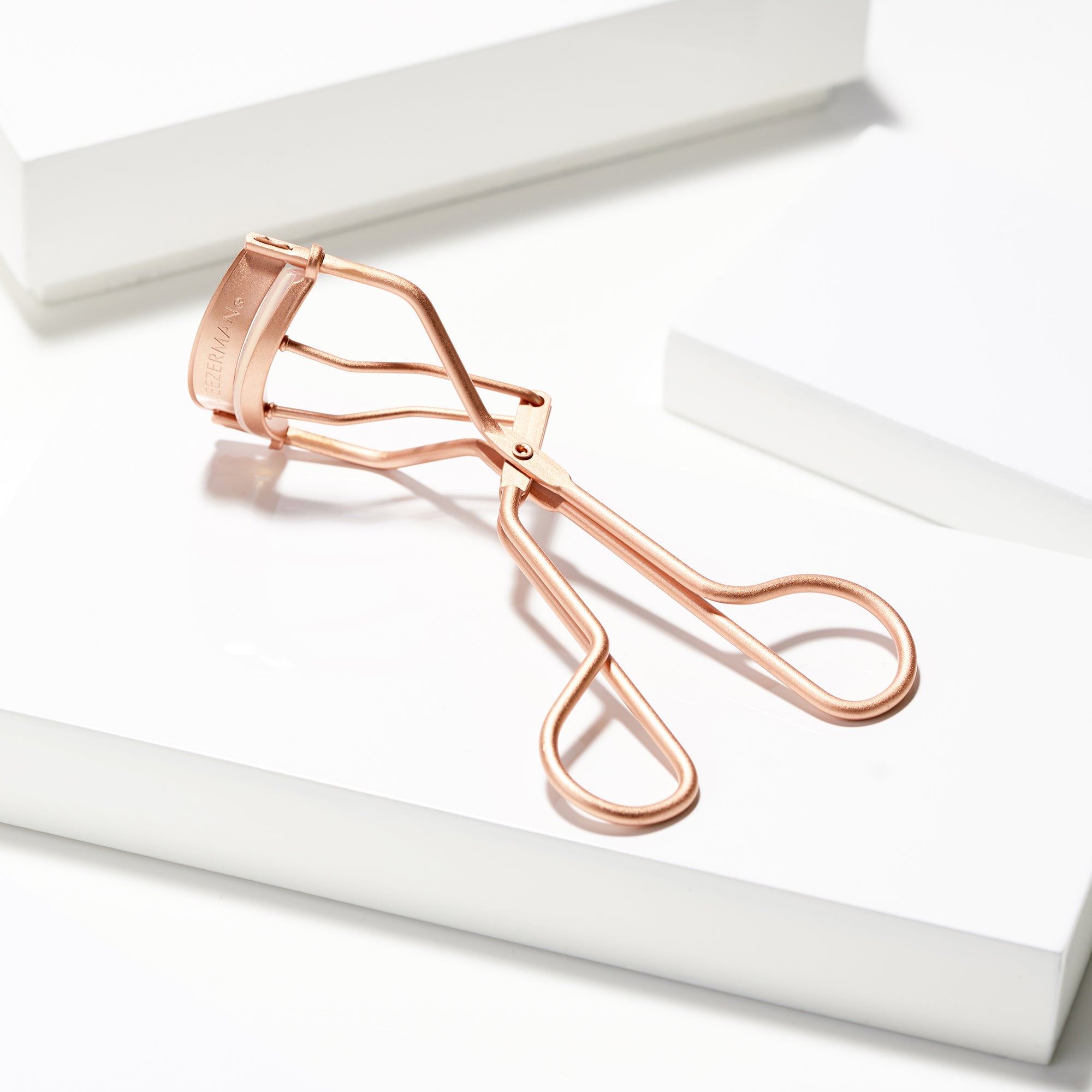 TWEEZERMAN klassiche Wimpernzange in der Farbe rose gold auf weißen Podesten.