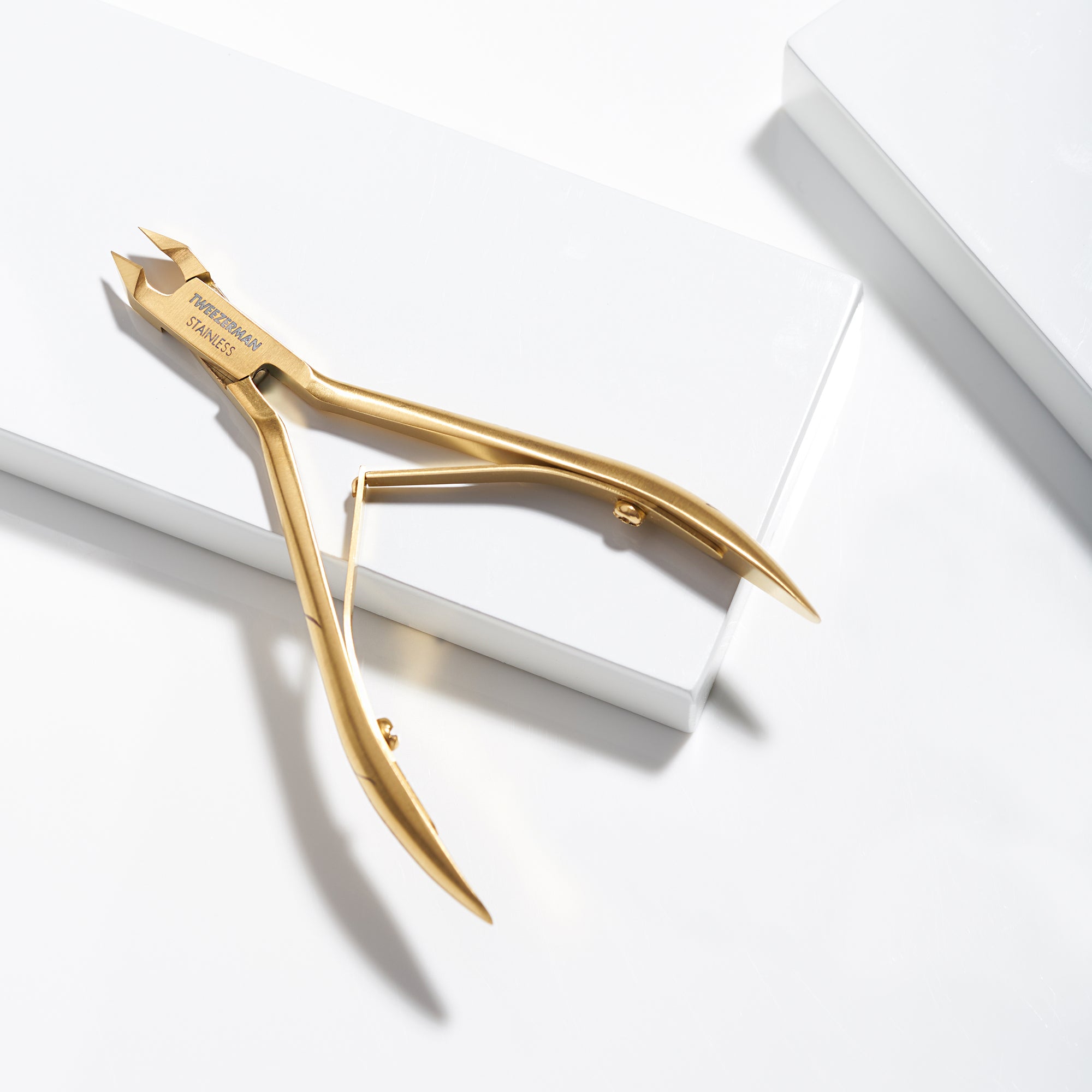 TWEEZERMAN Ultra Precision Nagelhautzange in gold aus rostfreiem Edelstahl auf weißen Podesten.