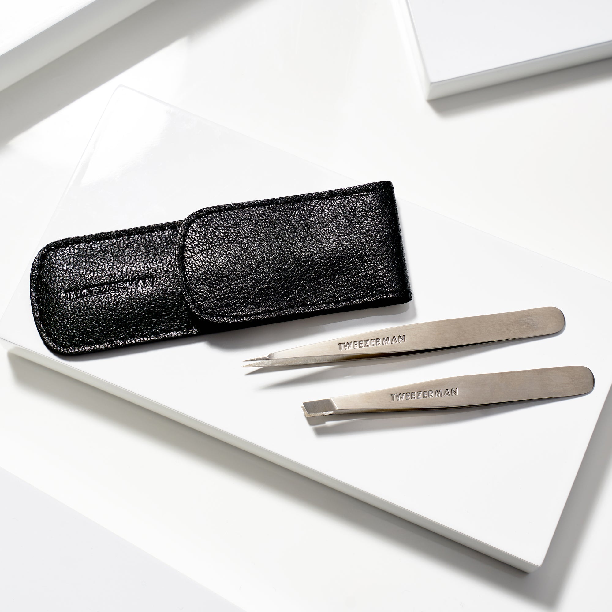 TWEEZERMAN Petite Set mit schräger und spitzer Pinzette in einem schwarzen Etui liegend auf weißen Podesten.