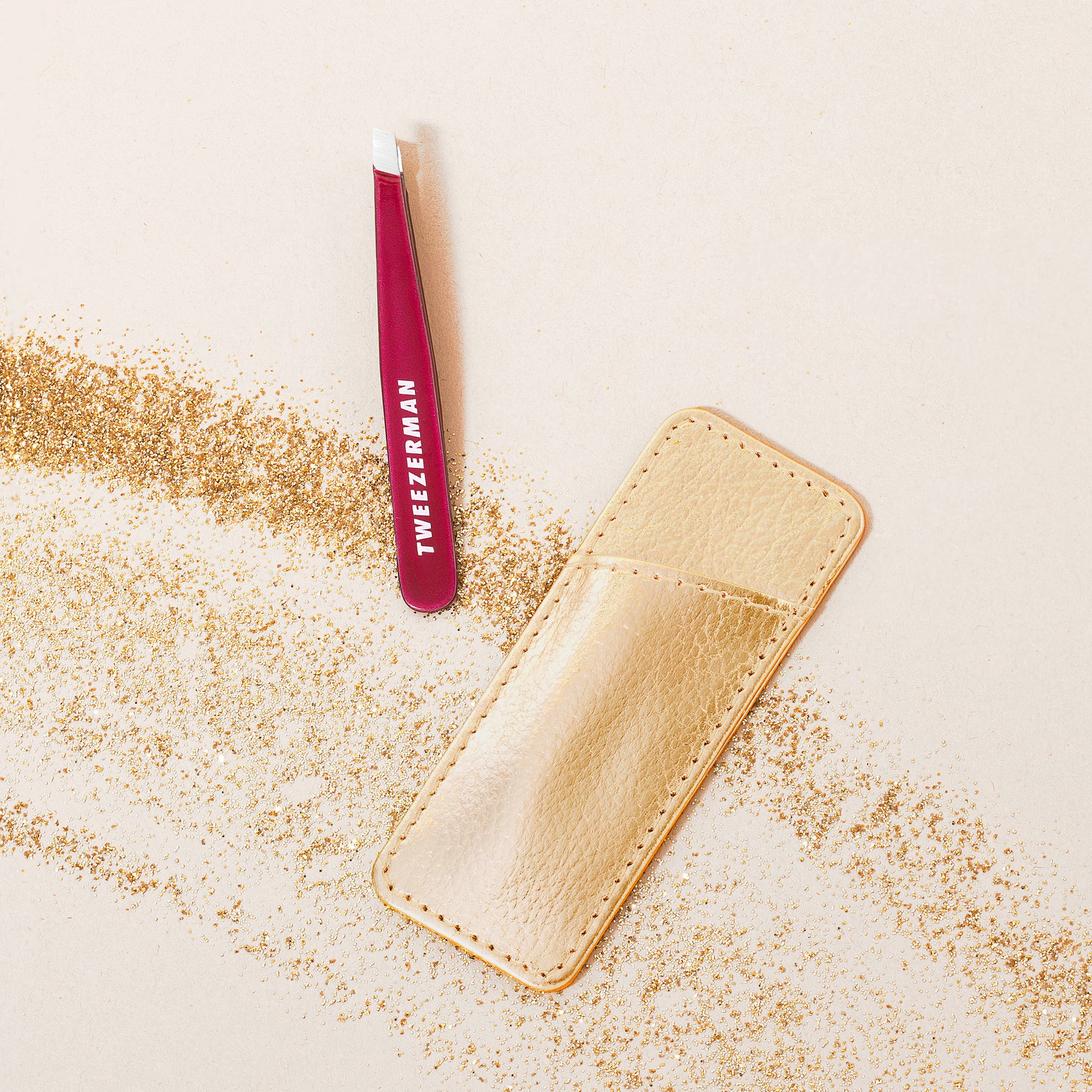 TWEEZERMAN Mini schräge Pinzette in fuchsia mit Etui umgeben von kleinen goldenen Steinchen.