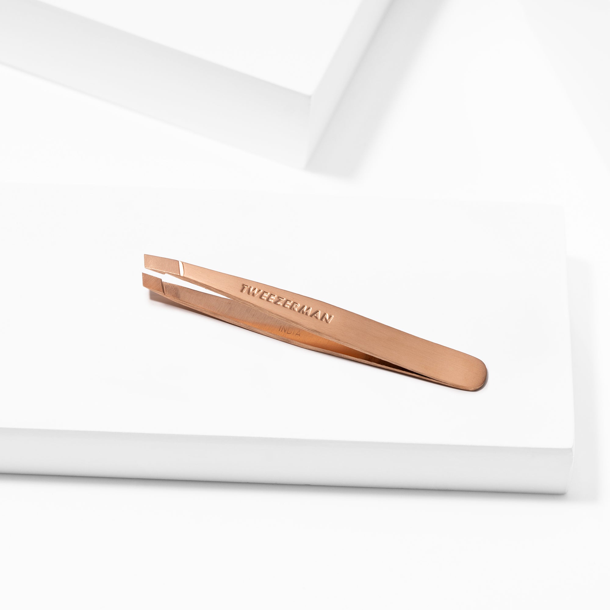 TWEEZERMAN Mini schräge Pinzette in rose gold liegend auf weißen Podesten.