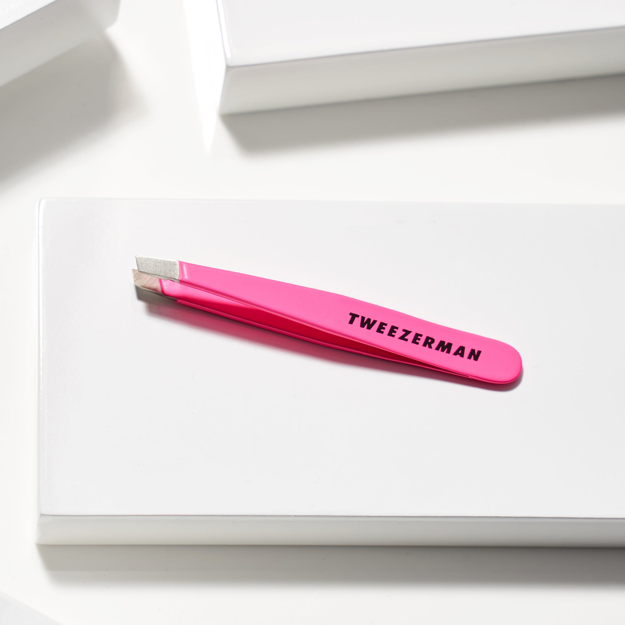 TWEEZERMAN Mini schräge Pinzette in neon pink liegend auf weißen Podesten.