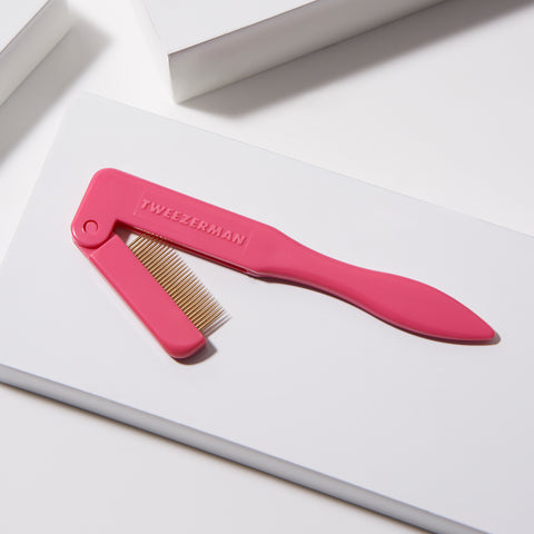 TWEEZERMAN faltbarer Wimpernkamm in pink mit vergoldeten Metallzinken auf weißen Podesten.