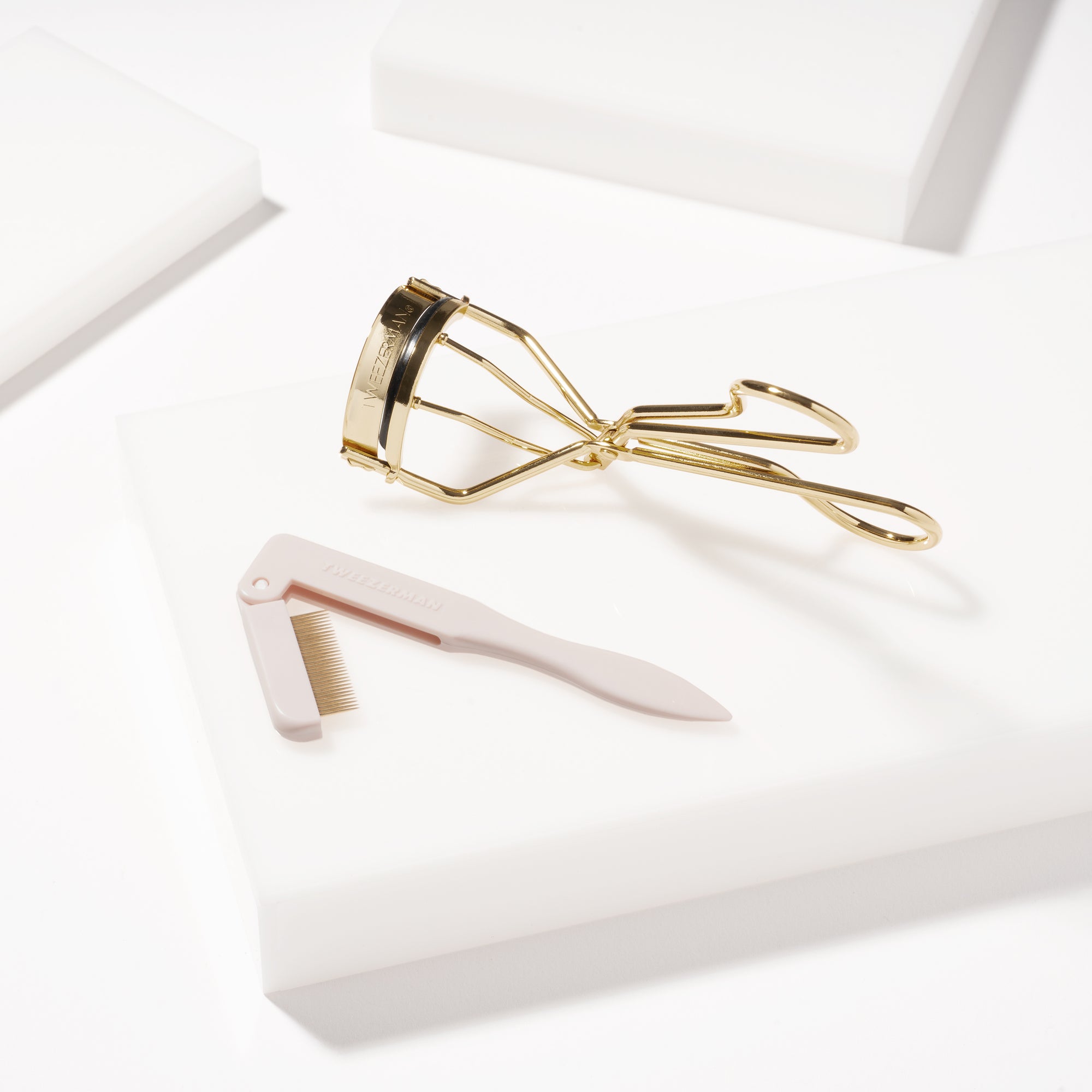 TWEEZERMAN Set mit klassischer Wimpernzange und Wimpernkamm in beige auf weißen Podesten.