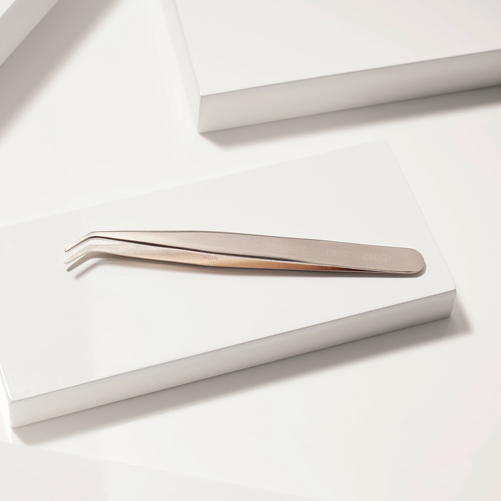 TWEEZERMAN Wimpernapplikator in rose Gold aus rostfreiem Edelstahl auf weißen Podesten.