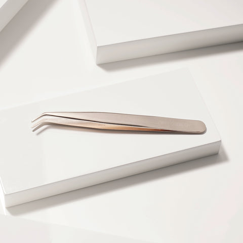 TWEEZERMAN Wimpernapplikator in rose Gold aus rostfreiem Edelstahl auf weißen Podesten.
