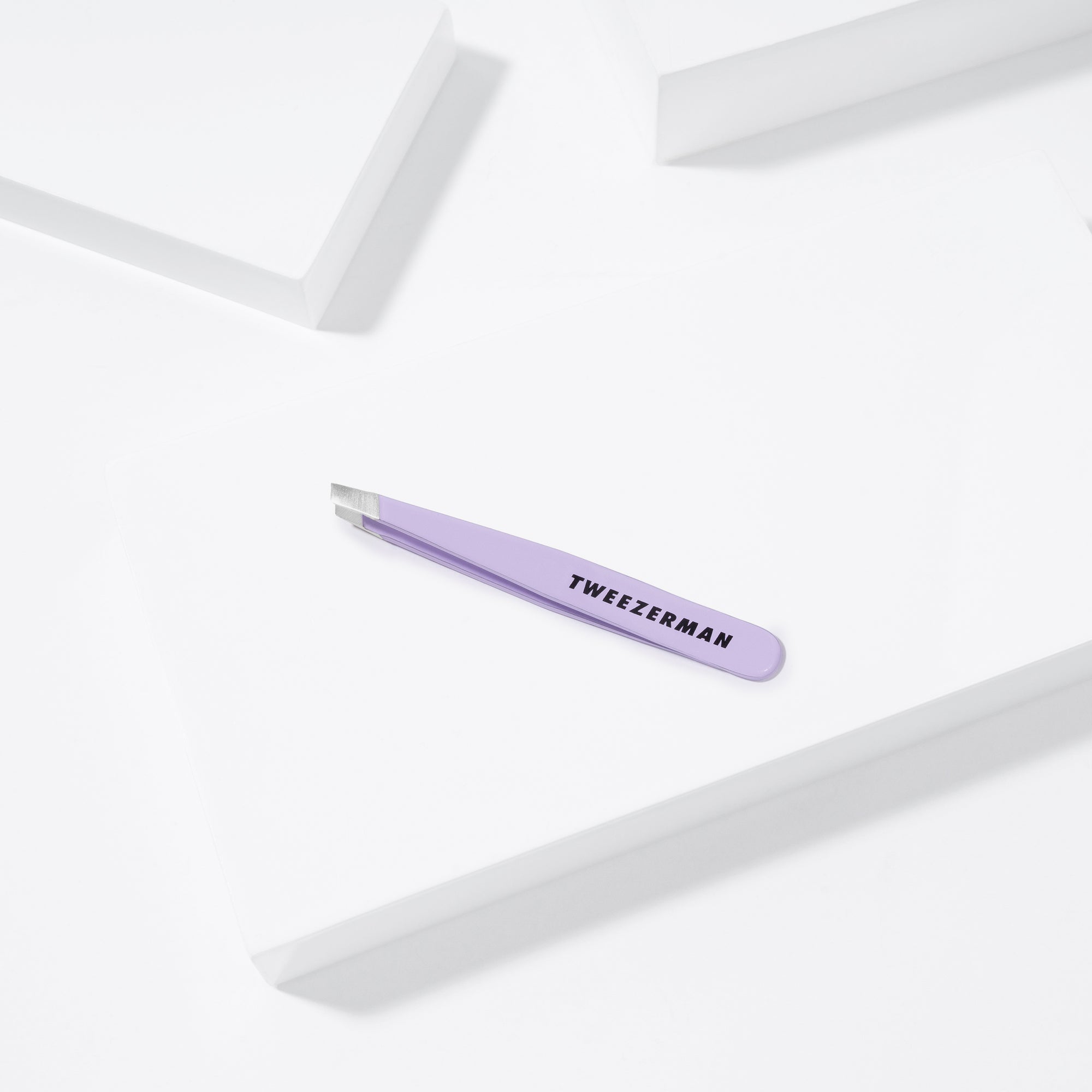 TWEEZERMAN Mini schräge Pinzette in lavender liegend auf weißen Podesten.