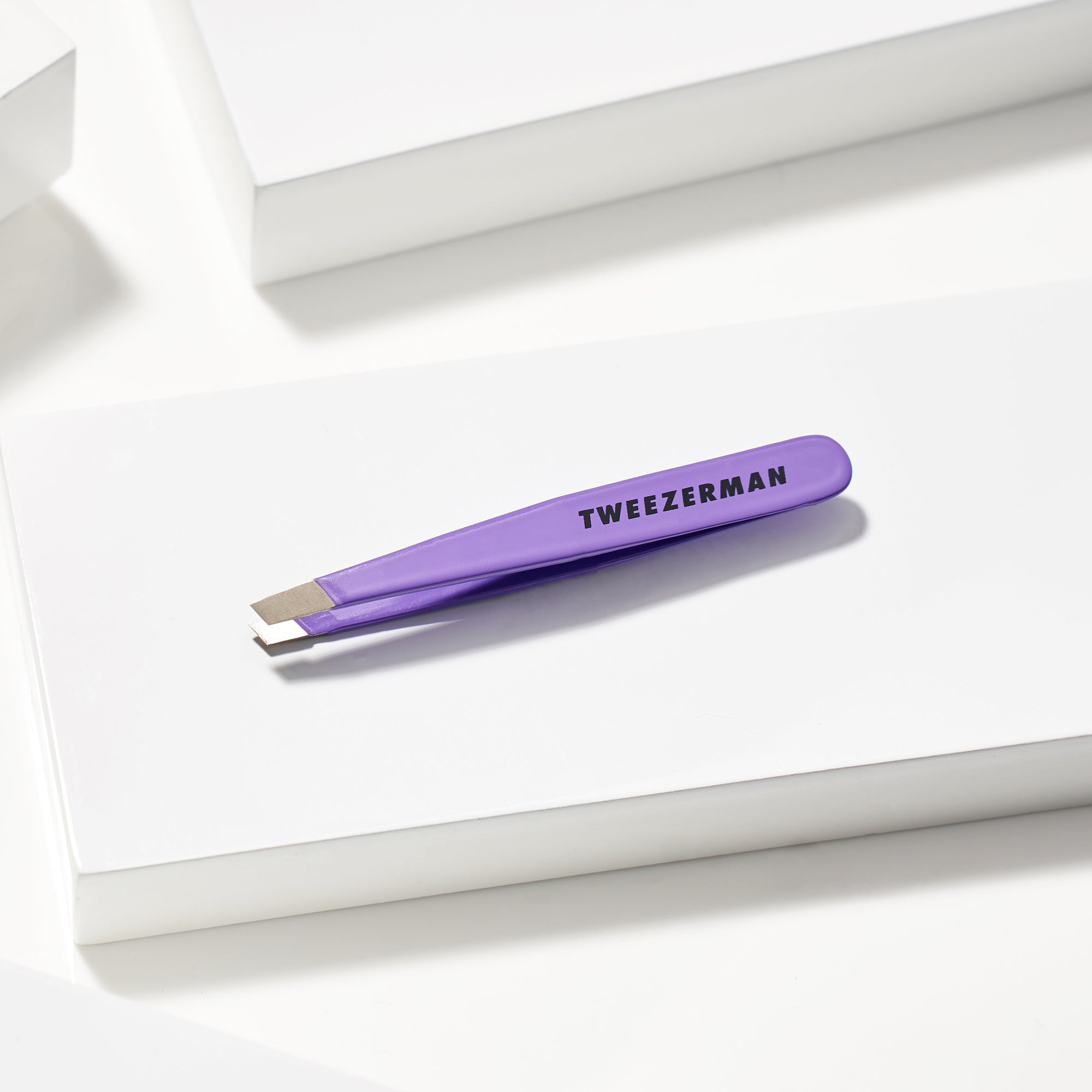 TWEEZERMAN Mini schräge Pinzette in lila liegend auf weißen Podesten.