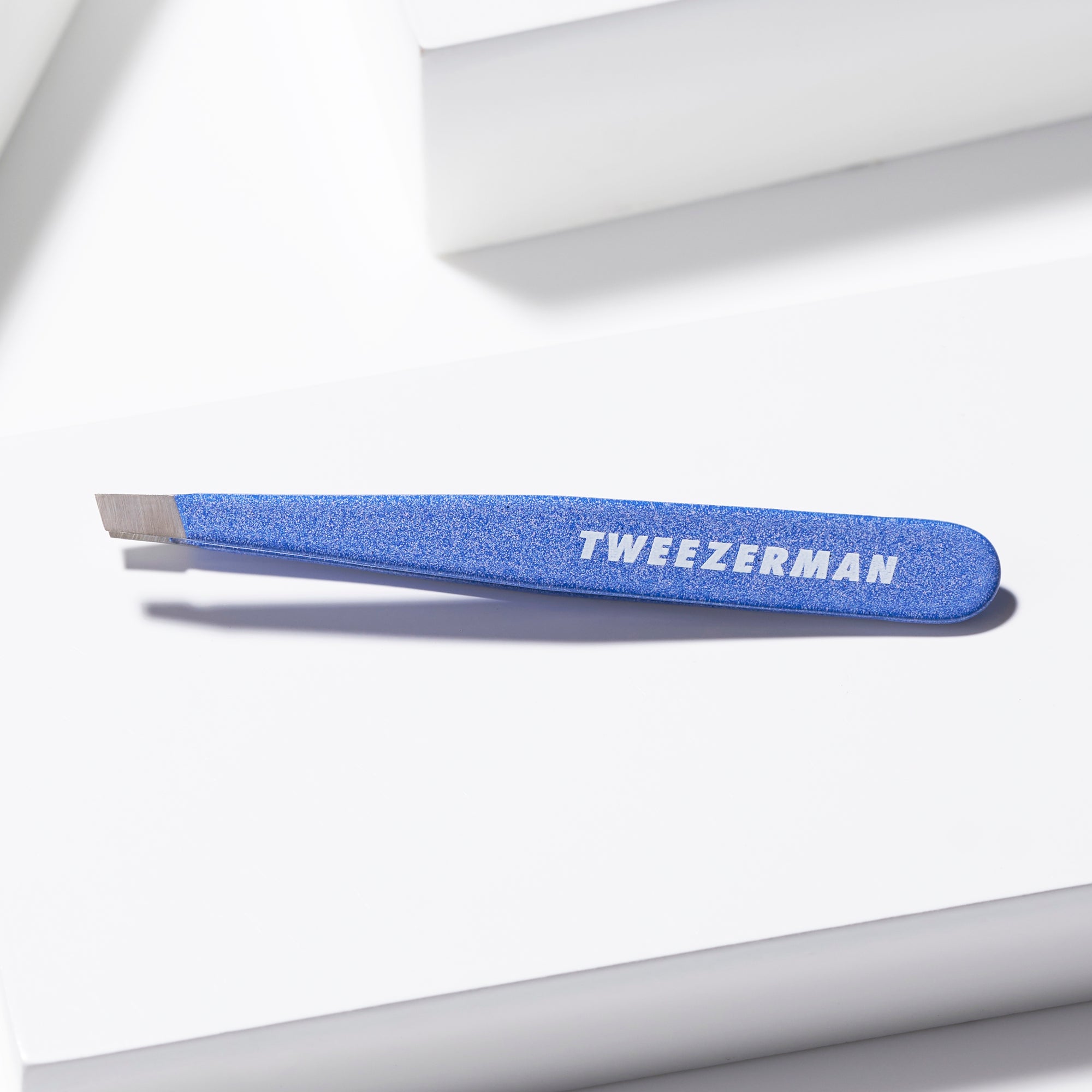 TWEEZERMAN Mini schräge Pinzette in blau liegend auf weißen Podesten.