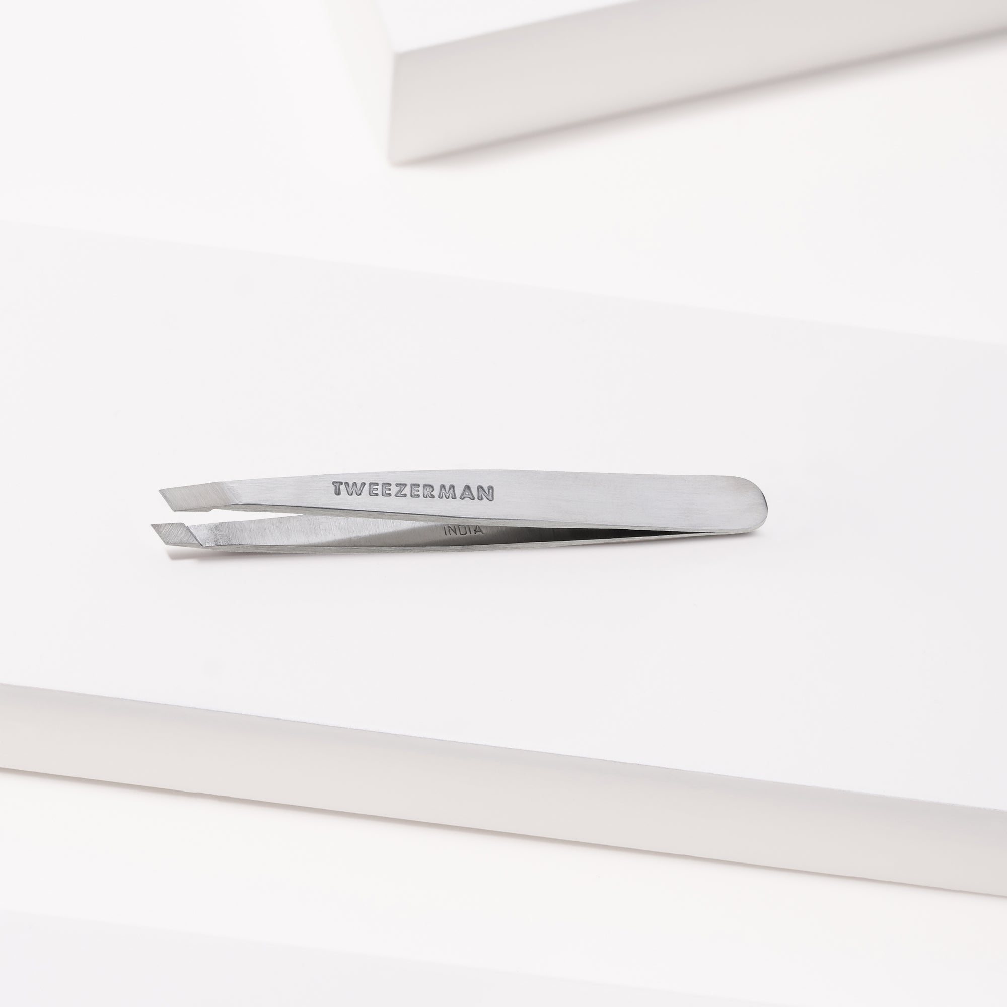 TWEEZERMAN Mini schräge Pinzette in silber liegend auf weißen Podesten.