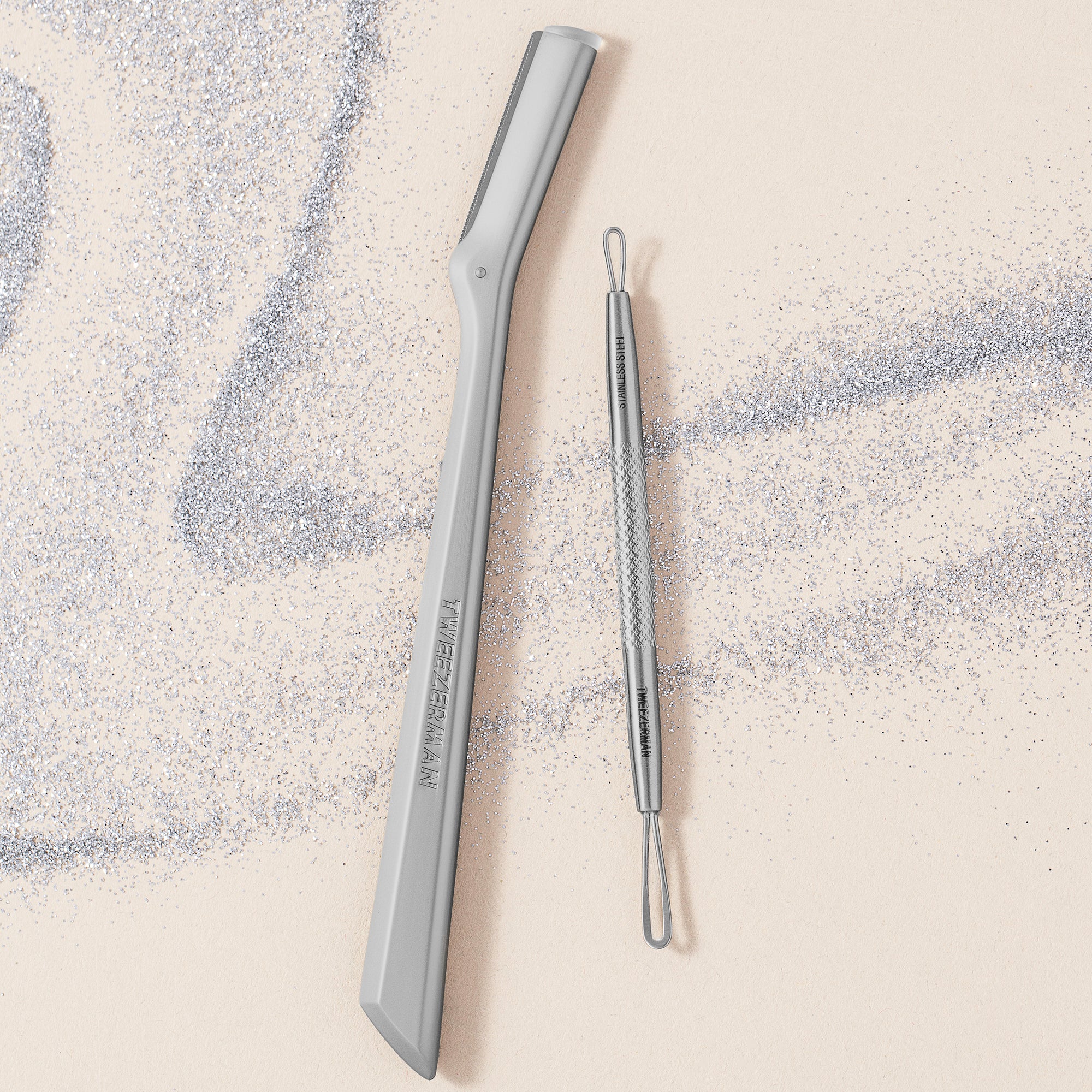 TWEEZERMAN Geschenkset mit dem Gesichtshaarrasierer und Mitesserentferner in silber aus rostfreiem Edelstahl umgeben von Glitzer.