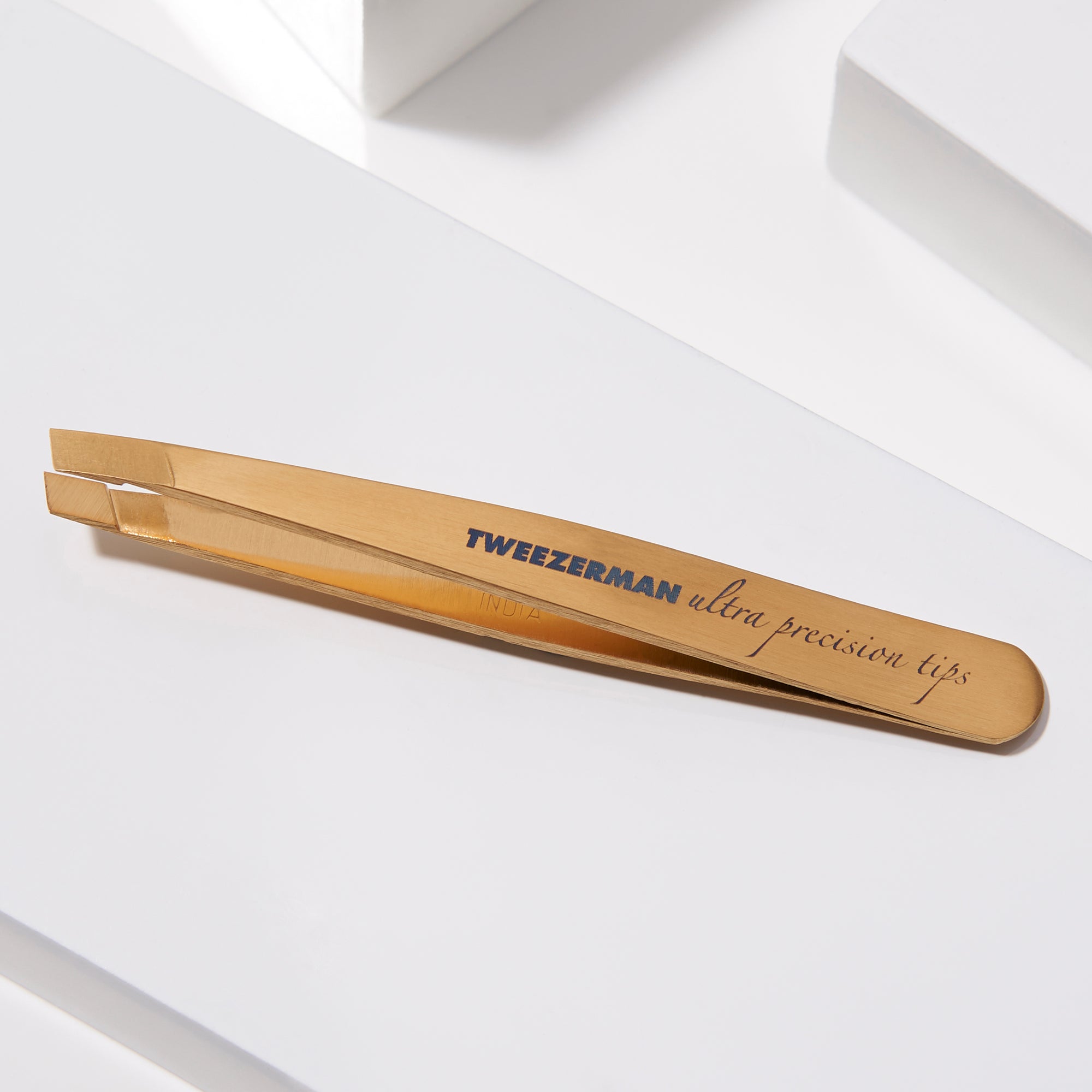 TWEEZERMAN Mini schräge Pinzette in gold liegend auf weißen Podesten.
