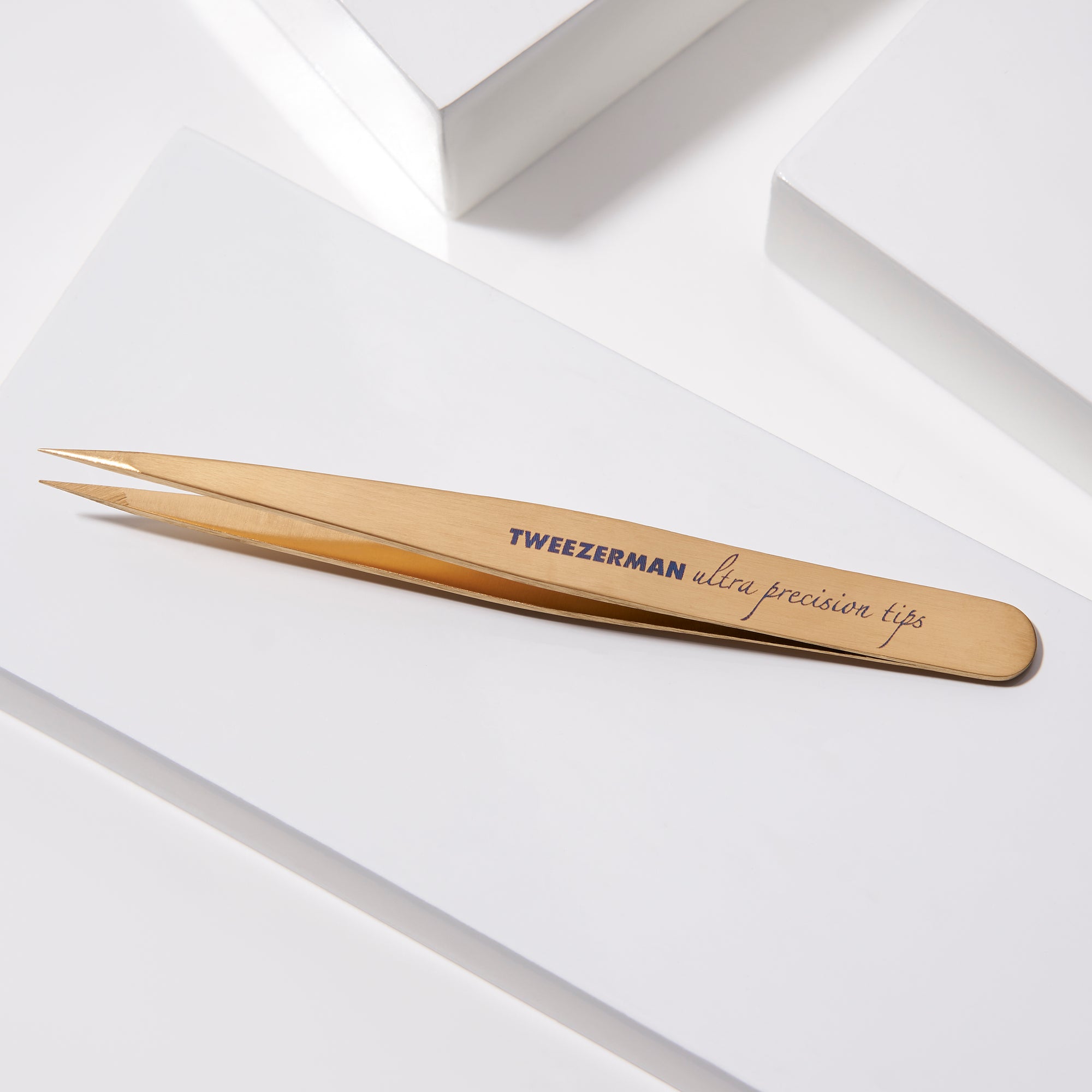 TWEEZERMAN spitze Pinzette in gold liegend auf weißen Podesten.
